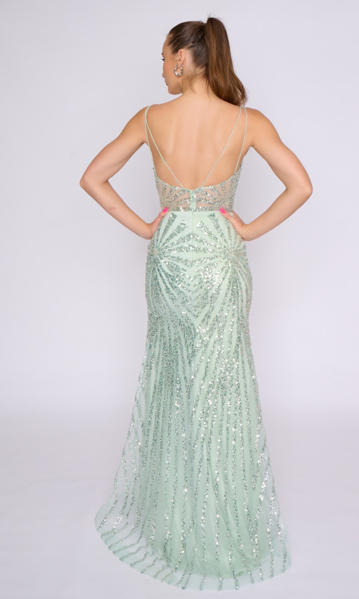 A woman stands with her back to the camera in a light green Nina Canacci 3267, a sleeveless long formal dress featuring floor-length sequins and a low back.